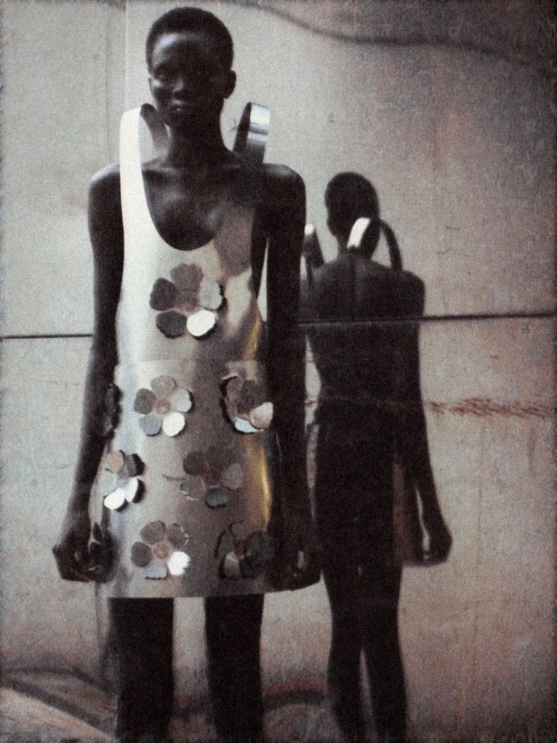 A slender, dark-skinned model in an avant-garde, shiny metal mini-dress with flower appliques poses against a mottled grey wall indoors.