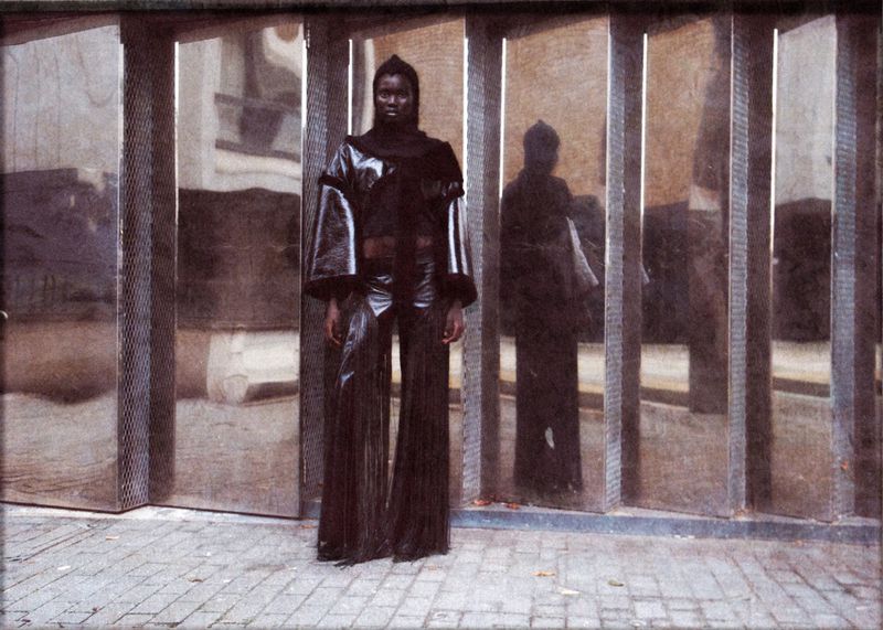 An adult female model with dark skin poses in a shiny dark outfit in front of a mirrored wall during the daytime in an urban area.