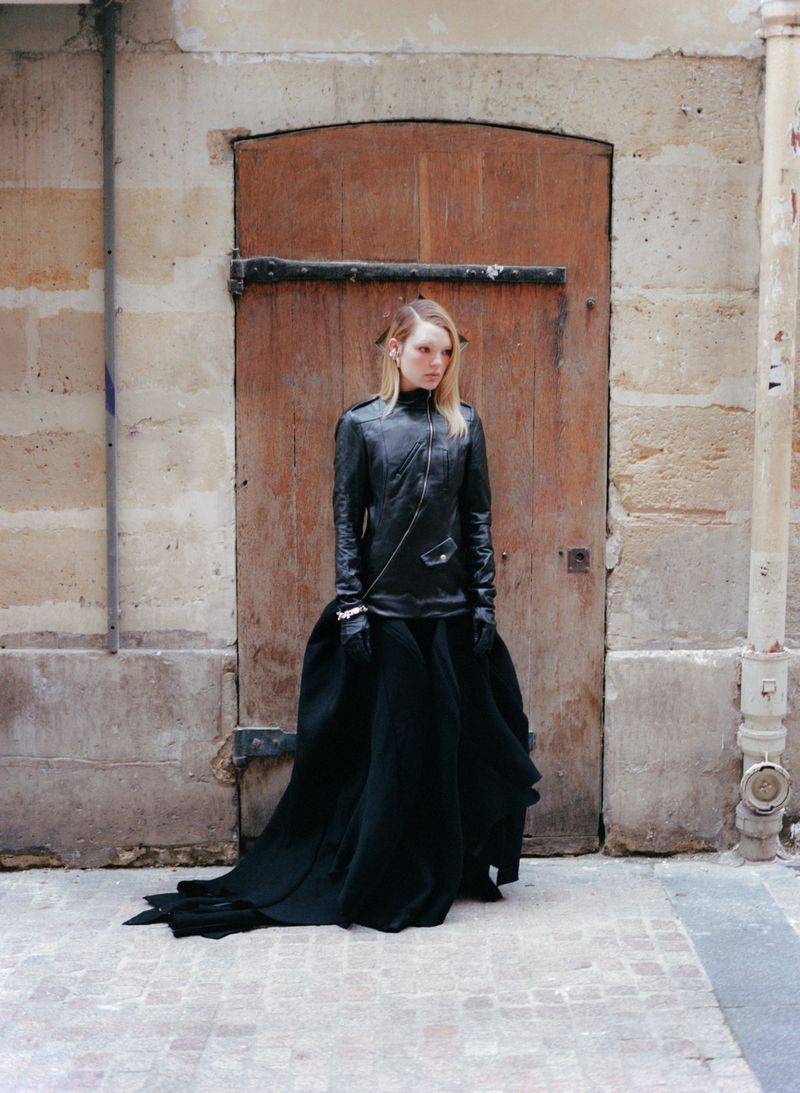 A slender woman stands in a black leather jacket and a long black skirt against a tan stone wall and a weathered brown door in an urban environment.