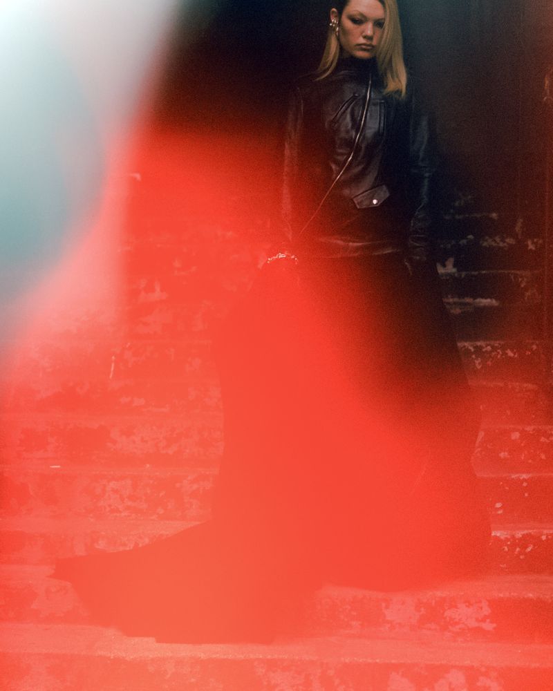 An adult female stands on concrete steps in black leather jacket, with intentional soft focus and muted colors in an outdoor shot.