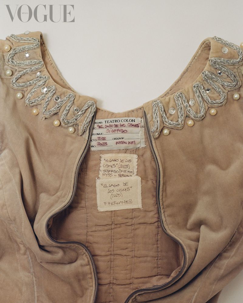 Close-up of a vintage beige velvet vest with pearl and sequin trim, adorned with handwritten tags, and the Vogue logo in the upper left.