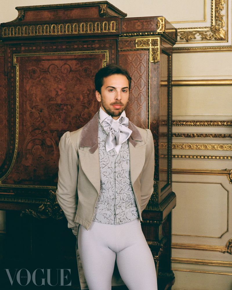 A portrait of a stylish adult male in a Regency-era suit, posing indoors with neutral expression, standing next to a dark wood cabinet.
