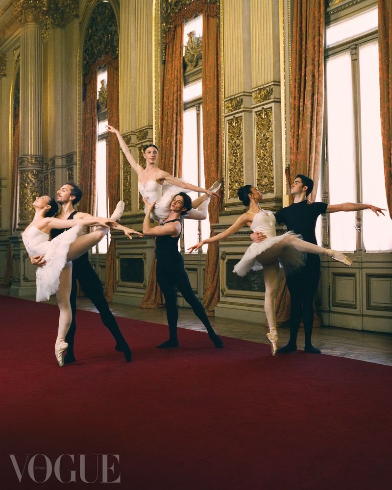 Ballet dancers in tutus and pointe shoes are lifted and posed by male dancers in an ornate hall with tall windows and red carpet.