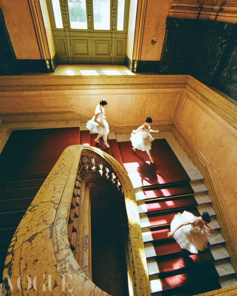 Three women in white dresses descend a grand marble staircase with red carpet in an overhead shot highlighting architecture and fashion.