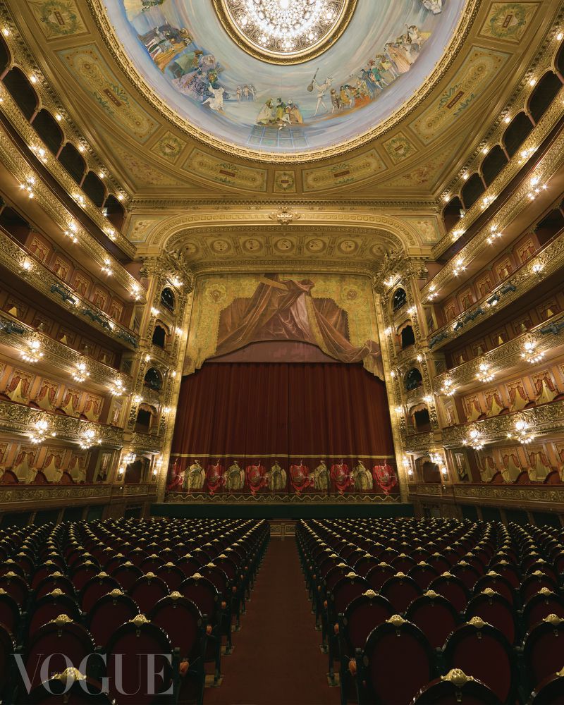 Ornate theatre interior featuring rows of red velvet seats facing a stage with a closed curtain, under a decorative domed ceiling.