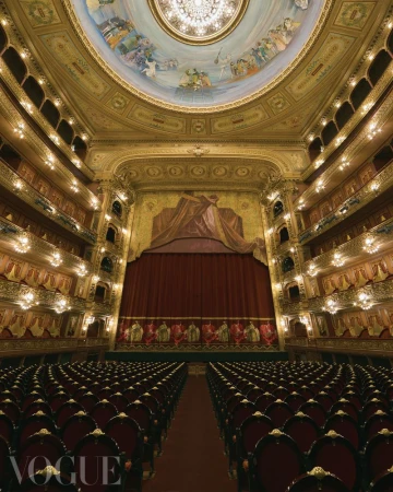 Ornate theatre interior featuring rows of red velvet seats facing a stage with a closed curtain, under a decorative domed ceiling.