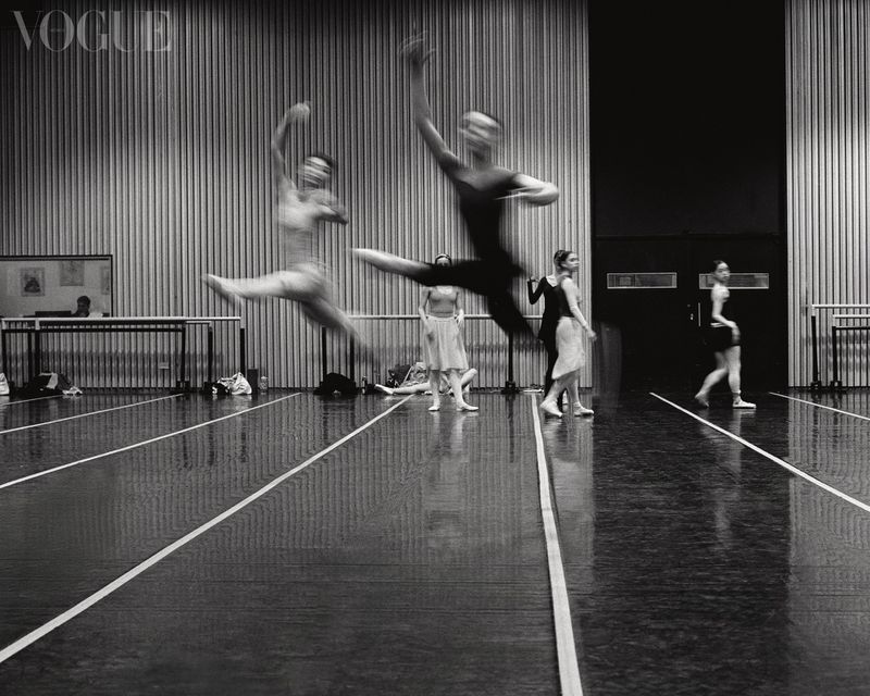 Black and white photo of ballet dancers leaping mid-air in a dance studio, captured during a performance, with a Vogue logo overlay.