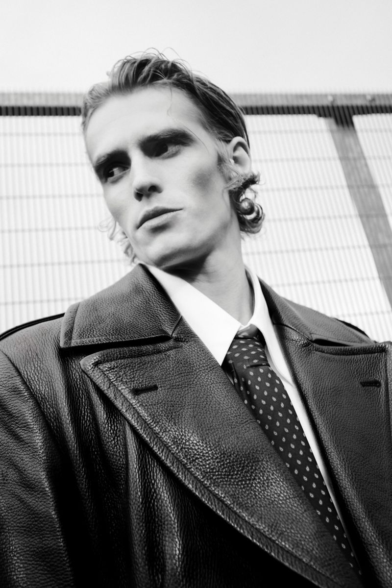 Black and white portrait of a man in a dark leather coat, white shirt, and tie against a light, geometric background.