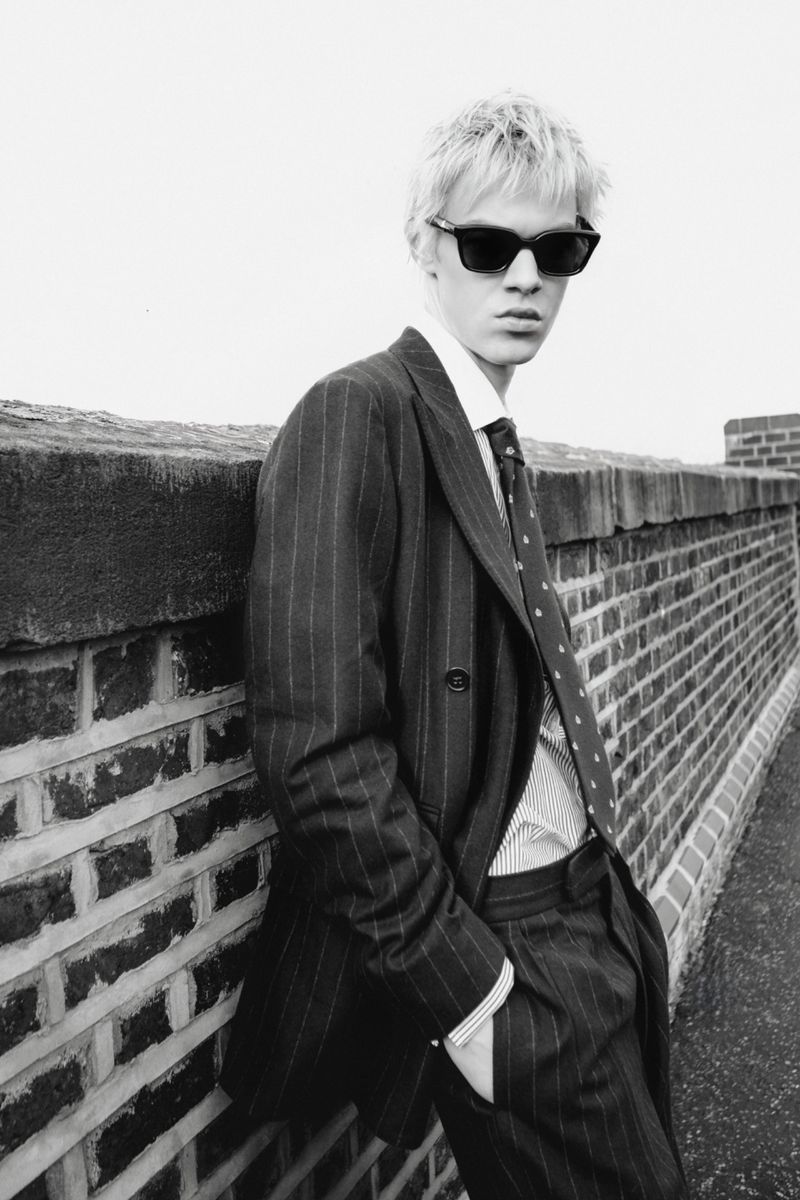 Young man in dark pinstripe suit and sunglasses leaning against a brick wall for Numéro Switzerland