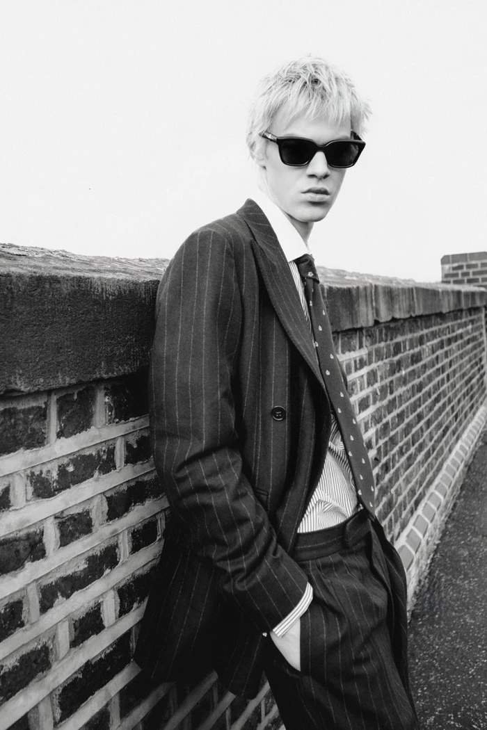Young man in dark pinstripe suit and sunglasses leaning against a brick wall for Numéro Switzerland