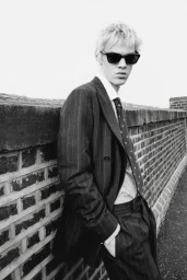 Young man in dark pinstripe suit and sunglasses leaning against a brick wall for Numéro Switzerland