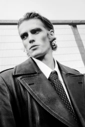 Monochrome portrait of a man looking upward wearing a textured black leather coat, white collared shirt, and patterned necktie.