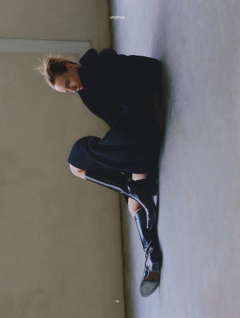Woman in a dark hooded jacket and patent leather boots poses against a neutral wall in a studio setting with soft, diffused lighting.