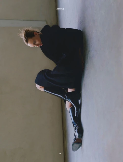 Woman in a dark hooded jacket and patent leather boots poses against a neutral wall in a studio setting with soft, diffused lighting.