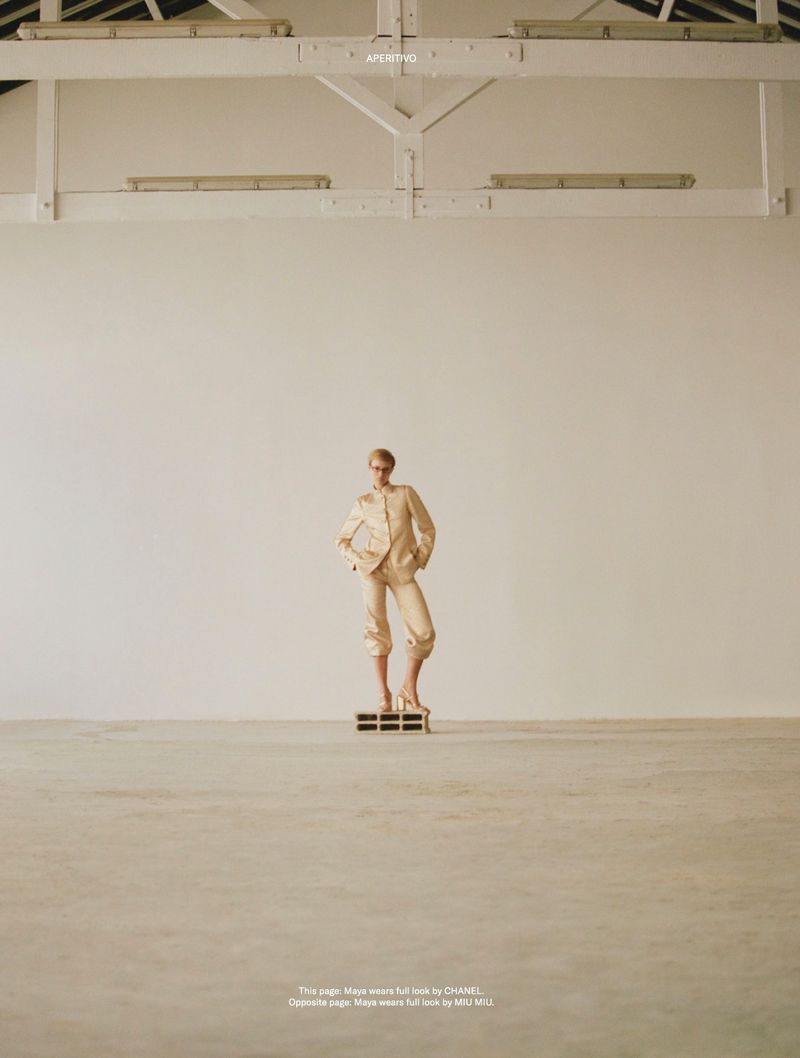 Fashion model in a beige suit posing on a platform in a light-filled studio with white walls and a high ceiling with exposed beams.