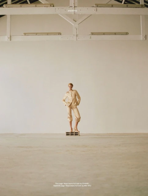 Fashion model in a beige suit posing on a platform in a light-filled studio with white walls and a high ceiling with exposed beams.
