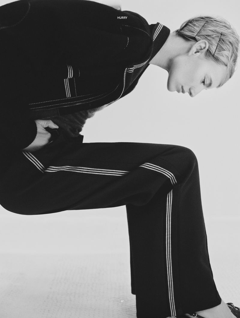 A woman in a black suit leans forward against a white background, her posture angled, creating a striking and modern fashion image.