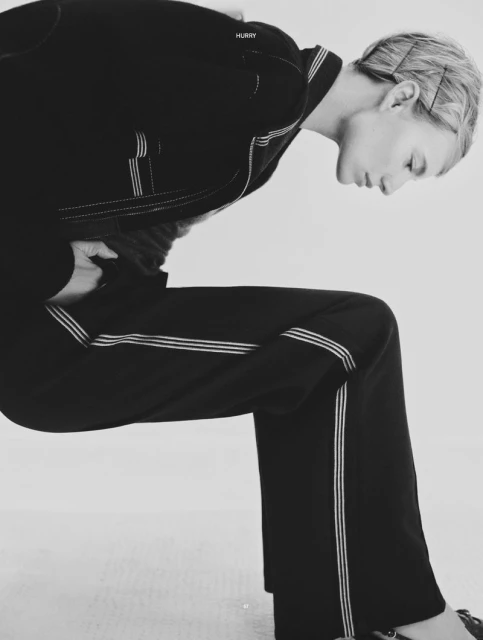 A woman in a black suit leans forward against a white background, her posture angled, creating a striking and modern fashion image.