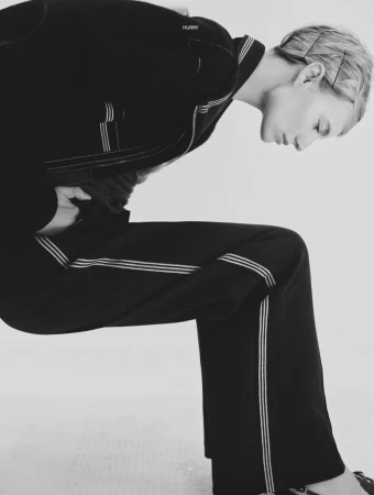 A woman in a black suit leans forward against a white background, her posture angled, creating a striking and modern fashion image.