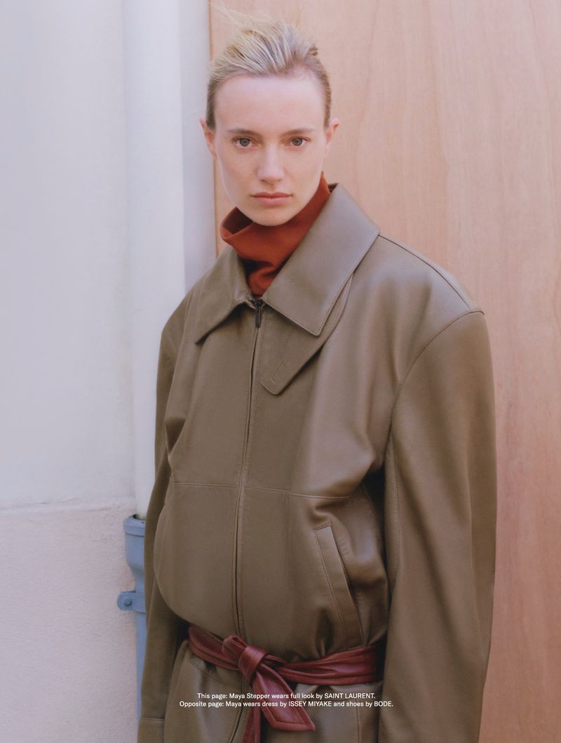 Fashion portrait of a blonde adult female wearing a leather jacket, brown turtleneck, and wine-colored belt, posing indoors against a light backdrop.