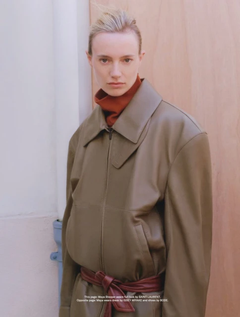 Fashion portrait of a blonde adult female wearing a leather jacket, brown turtleneck, and wine-colored belt, posing indoors against a light backdrop.