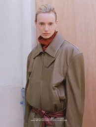 Model wearing an olive green leather coat and rust turtleneck posing against a pale wood wall for Family Style HURRY project.