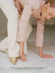 Model Maya Stepper wearing a pink jacket, cream trousers, and gold spiral earrings posing in a neutral studio interior.