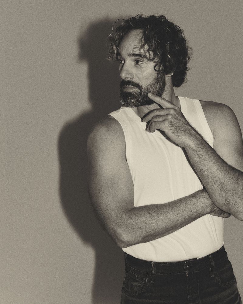 Monochrome portrait of adult male in white tank top against neutral background, gazing pensively to the side with hand to face.