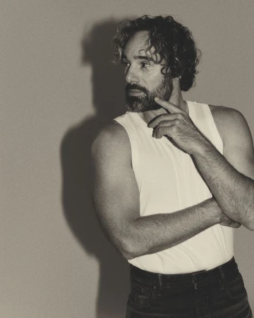 Monochrome portrait of adult male in white tank top against neutral background, gazing pensively to the side with hand to face.