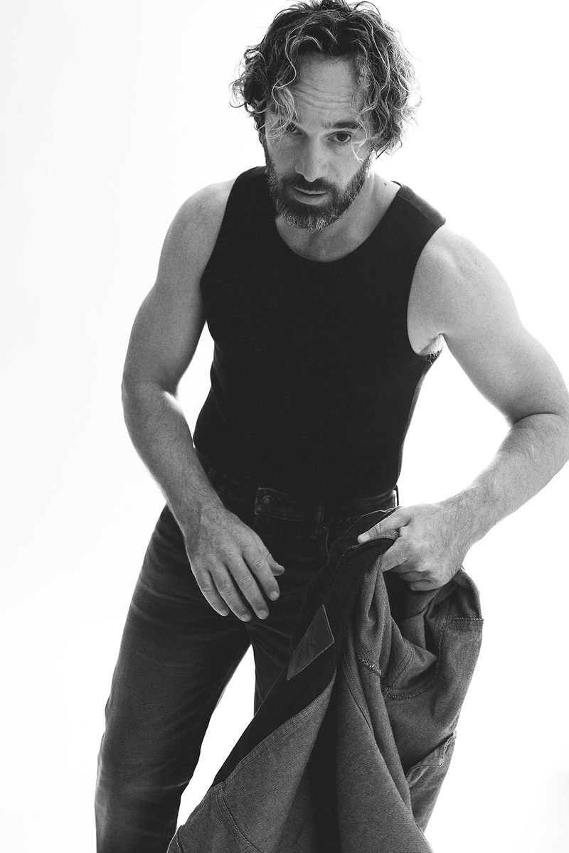 Black and white portrait of an adult male wearing a black tank top holding a denim jacket in a brightly lit photo studio.