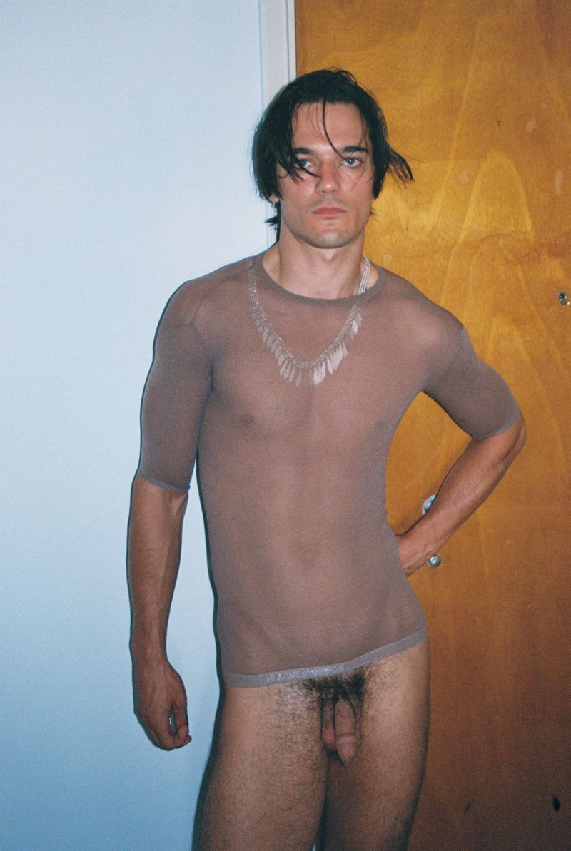 Portrait of a slim adult male, medium skin tone, posing with a sheer top, against a neutral door and wall in an indoor setting.