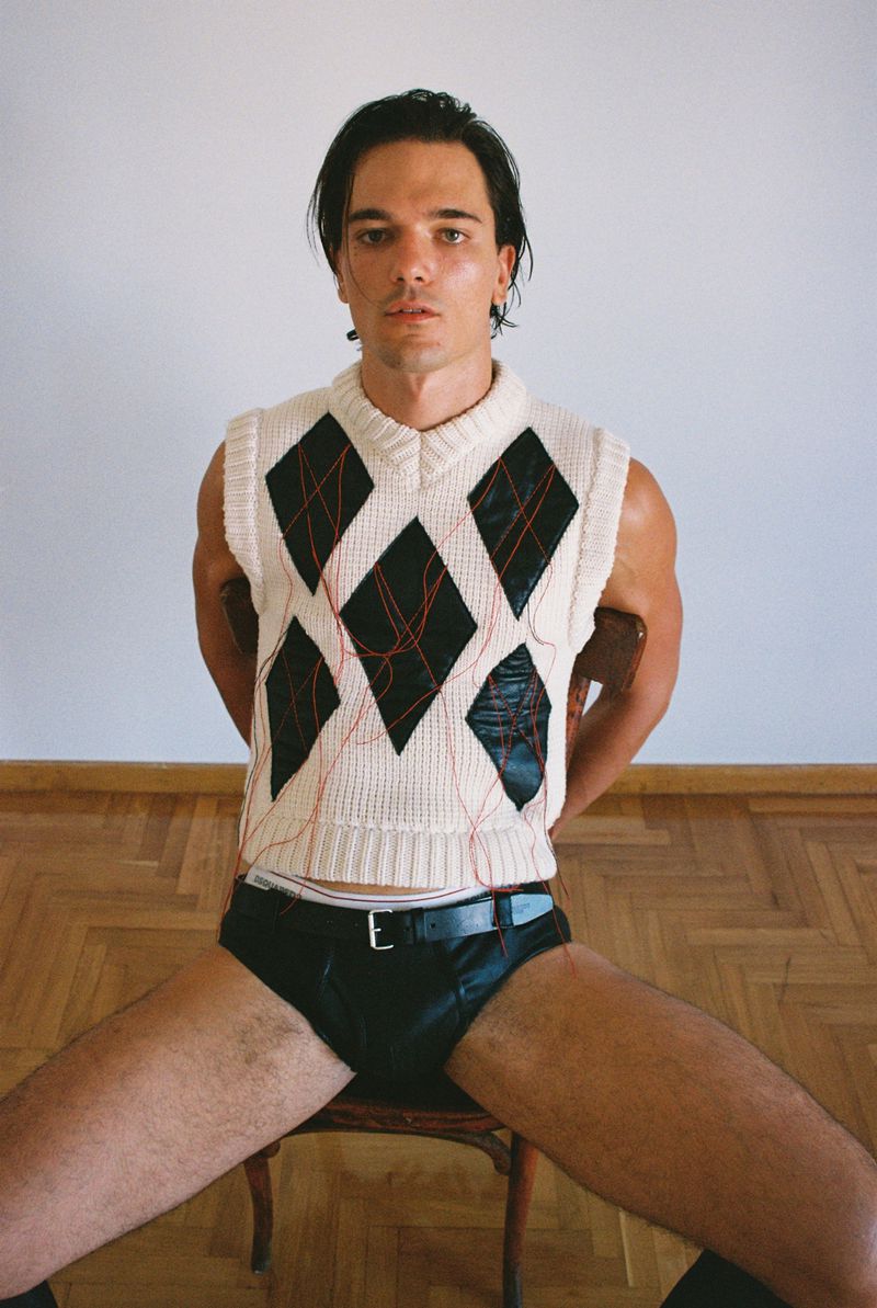 Portrait of a young man with dark hair in a vest and black briefs, seated on a wooden chair, posing in a minimalist white studio.
