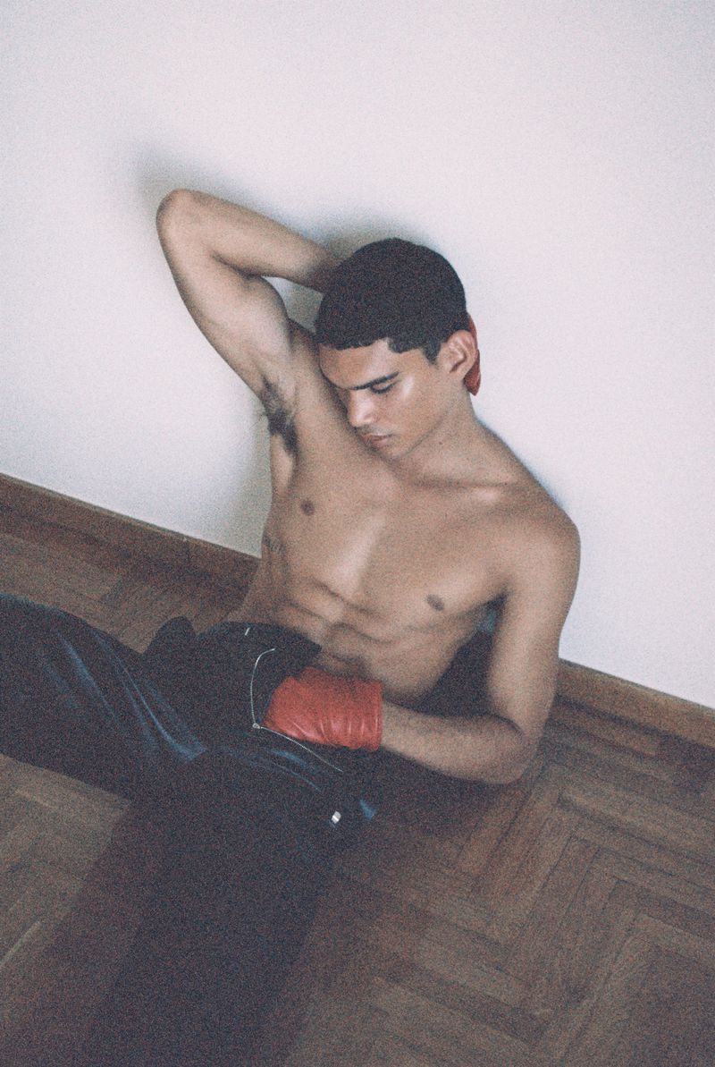 Athletic man poses indoors, shirtless with boxing gloves and pants. Light streams from above, creating a studio-like atmosphere.
