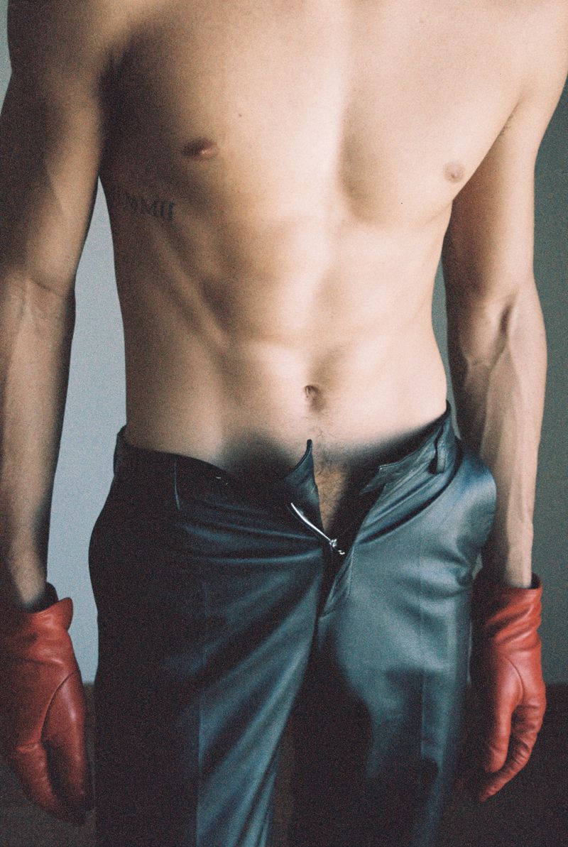 Torso of a muscular adult male with open dark pants and red leather gloves in a close-up shot against a plain background.