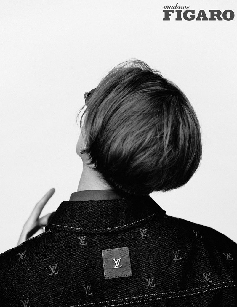 Black and white fashion shot of a person with dark hair wearing a dark jacket with an 'LV' pattern against a white background.