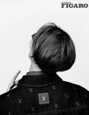 Black and white fashion shot of a person with dark hair wearing a dark jacket with an 'LV' pattern against a white background.