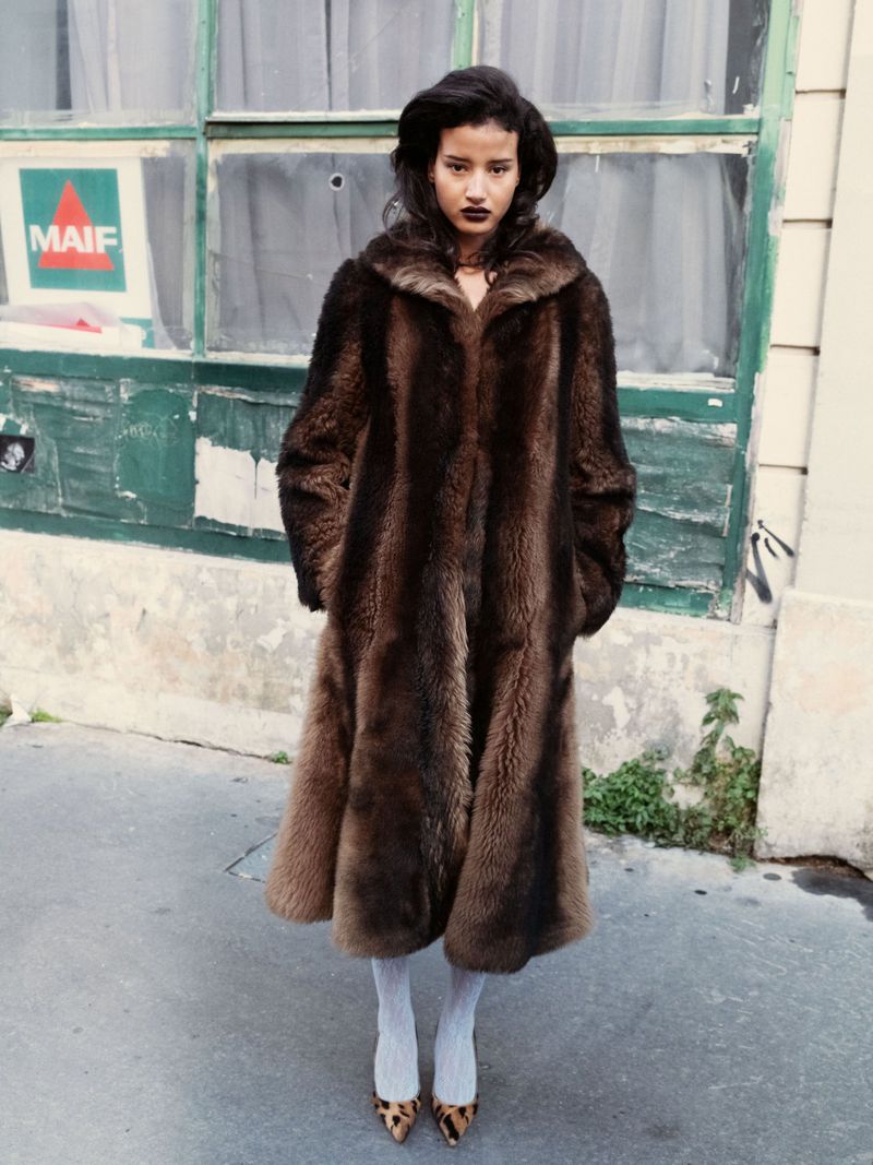A solo female adult stands on a street wearing a fur coat, tights, and shoes. The background features a green building facade.