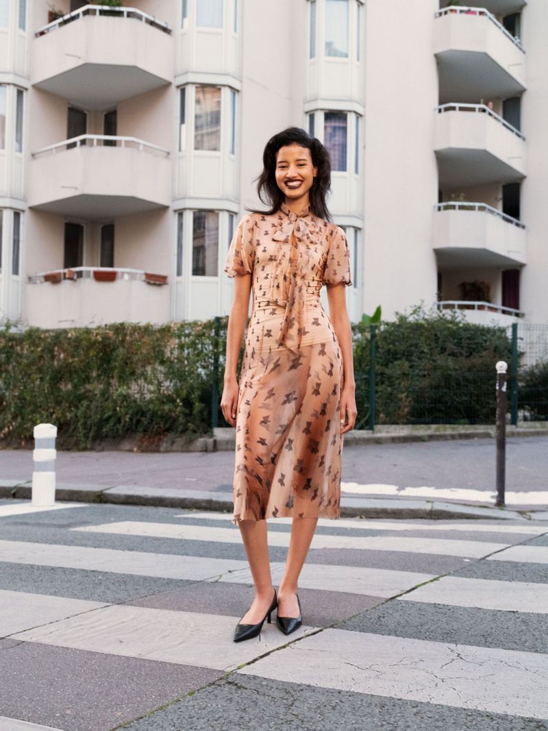 A woman in a peach-colored dress stands on a crosswalk in front of an apartment building, smiling and looking toward the viewer.