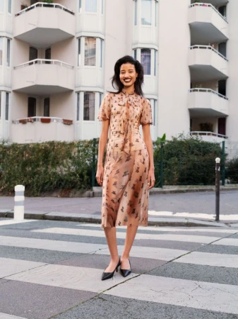 A woman in a peach-colored dress stands on a crosswalk in front of an apartment building, smiling and looking toward the viewer.
