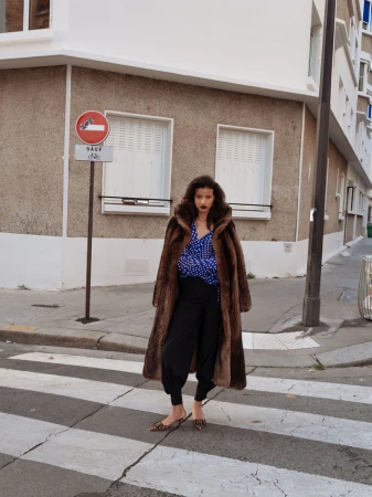 Woman in fur coat and black pants standing on crosswalk in urban setting with a no-entry sign in the background, fashion style.