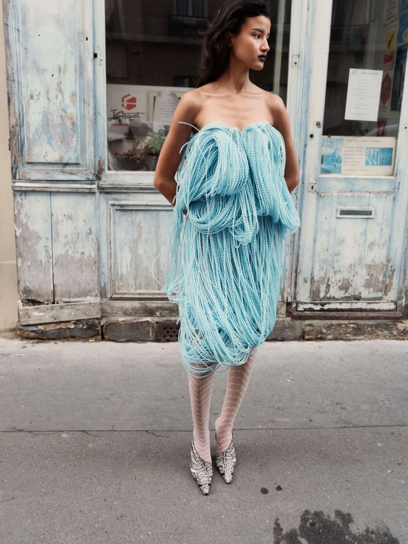 Fashion portrait of an adult woman wearing a light blue dress and decorated heels standing in front of a weathered blue wall.