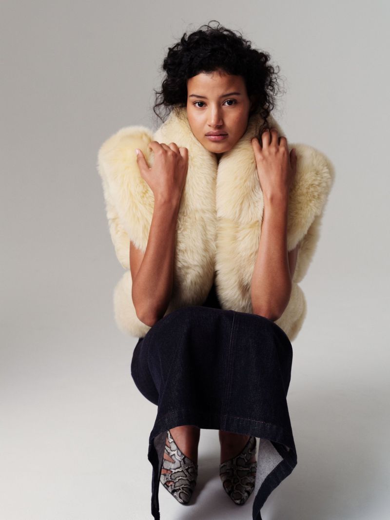 Adult female poses in a fur vest and patterned heels against a neutral backdrop, fashion photography, full shot.