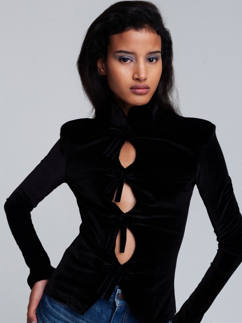 Adult female model poses in a black velvet long sleeve top with front cutouts and blue jeans against a neutral gray backdrop.
