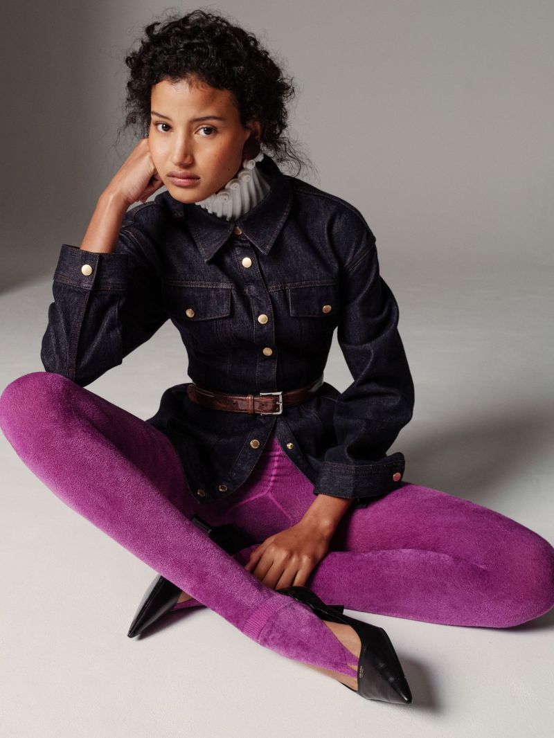 A young woman in a denim jacket and purple leggings poses while sitting on the floor in a studio environment.