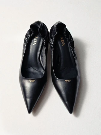 Two black leather Prada pointed-toe pumps displayed on a white surface, showcasing fashion footwear with visible logo detail.