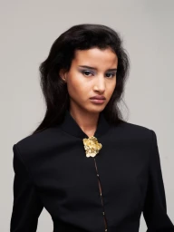 Model Khadija Lahmine in a black jacket with a gold floral brooch against a neutral grey studio background for Dazed MENA.