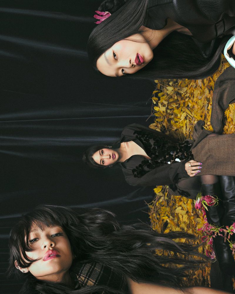 Three Asian women wearing dark, flowing clothing pose against a dark background with autumn foliage details in a fashion editorial.