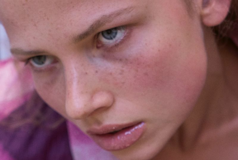 Close-up portrait of a fair-skinned young adult woman with freckles, blue eyes, and glossy lips, looking slightly downward.