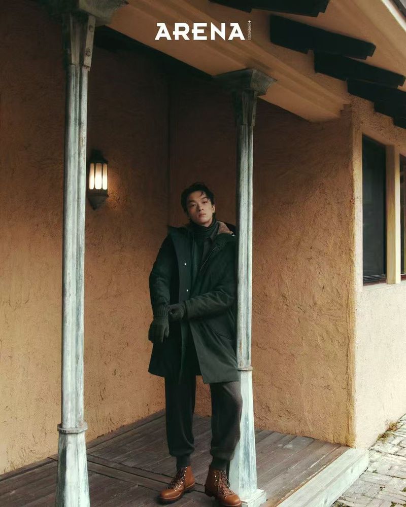 Man in olive coat and brown pants stands against building columns; outdoor fashion editorial with 'Arena' logo visible on top.