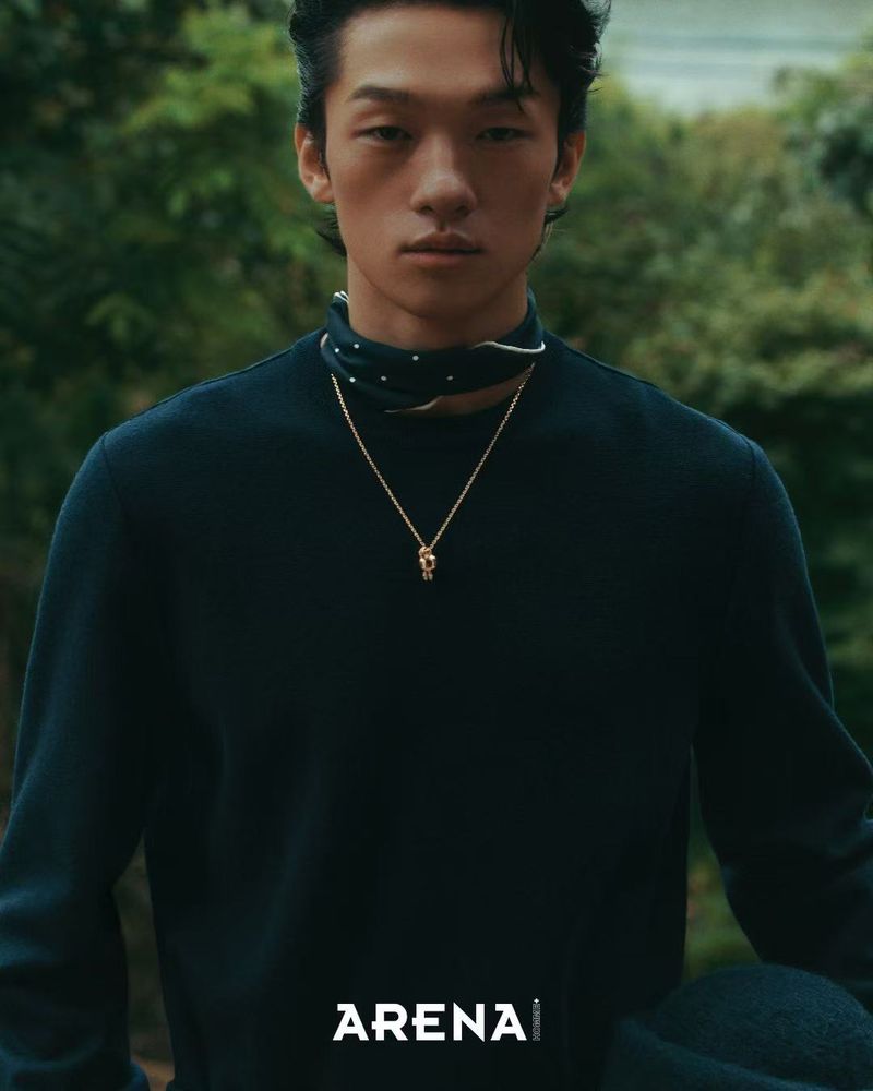 Portrait of an Asian male model with dark hair wearing a black top and gold necklace in an outdoor setting, looking at the camera.
